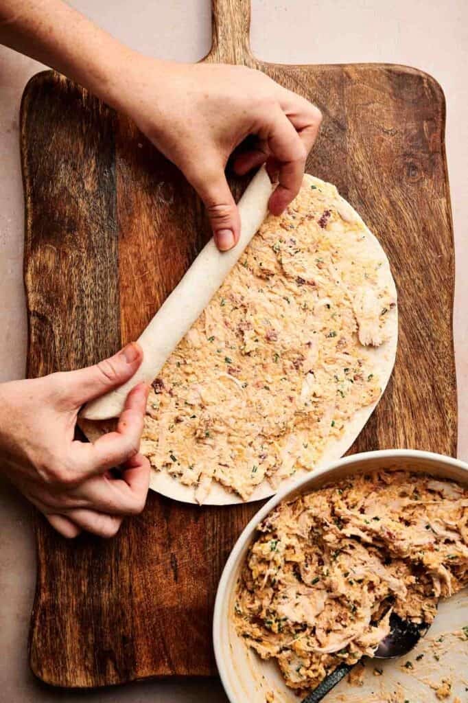 Hands rolling a tortilla filled with a creamy buffalo chicken pinwheel spread on a wooden cutting board; a bowl with extra filling sits nearby.