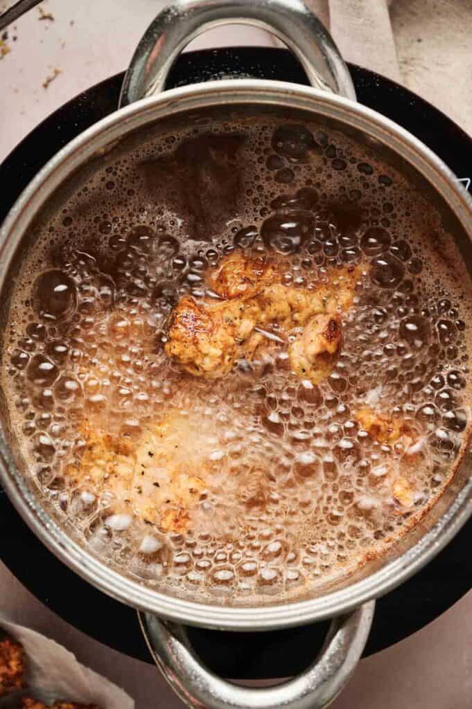Pieces of battered food, reminiscent of Chicken and Waffles, are being deep-fried in hot oil inside a metal pot, with bubbles rising around the food.