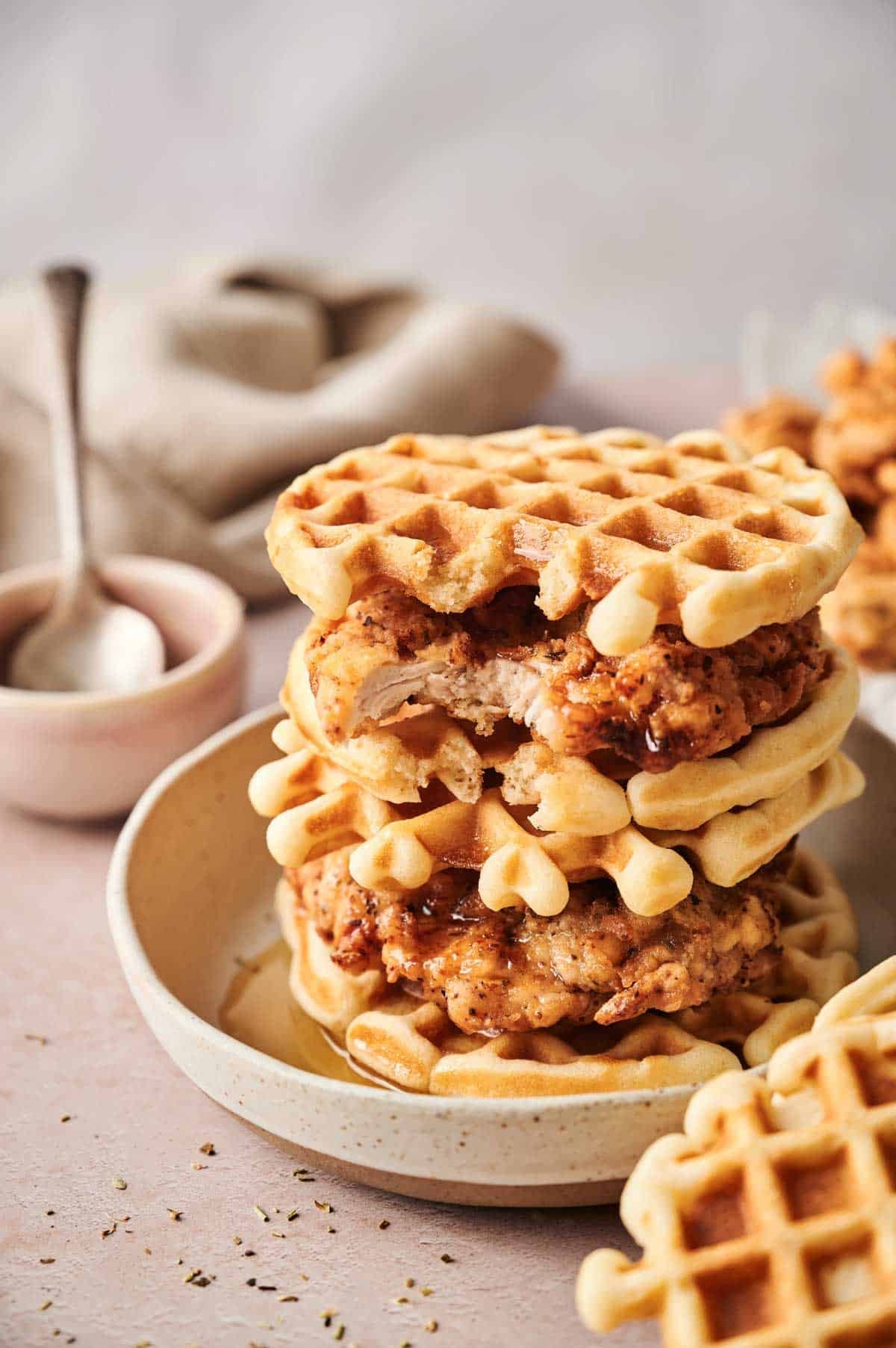 Three Chicken and Waffles sandwiches are stacked on a plate, syrup drizzled over each layer. In the background, a small bowl with a spoon and a napkin complete this delicious scene.