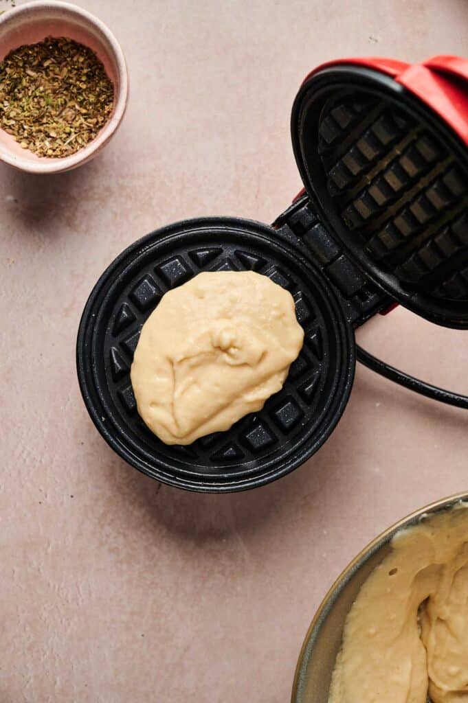 Waffle batter on a waffle maker next to a bowl of dried herbs and extra batter, perfect for serving with classic Chicken and Waffles on a light countertop.