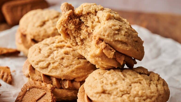 A close-up of sandwich cookies on parchment paper, with one cookie at the top missing a bite. Crumbs and broken cookie pieces are visible around them.