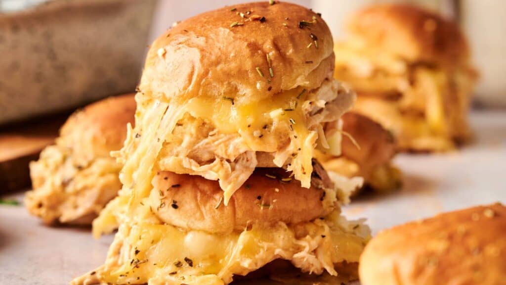 A close-up of stacked cheesy chicken sliders on soft buns, topped with herbs, with more sliders blurred in the background.
