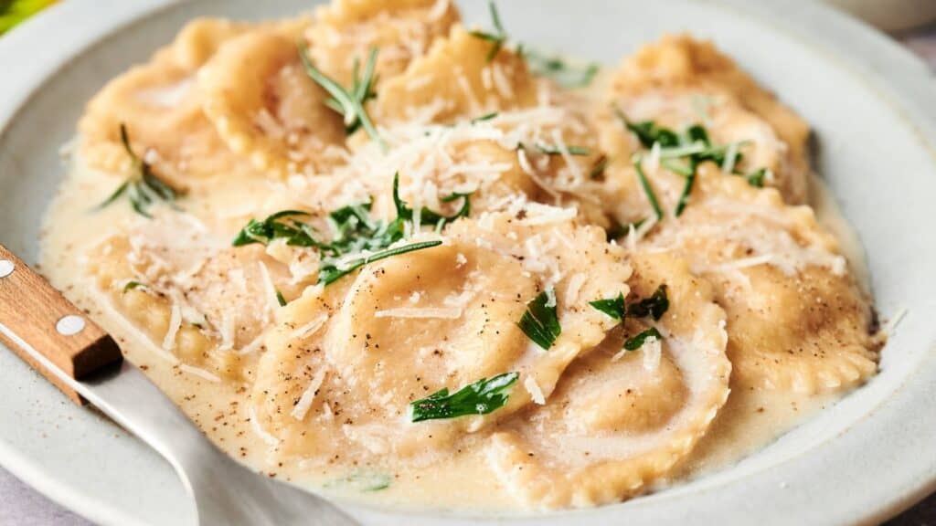 A plate of ravioli in a creamy sauce, garnished with grated cheese and fresh herbs, with a fork resting on the side.