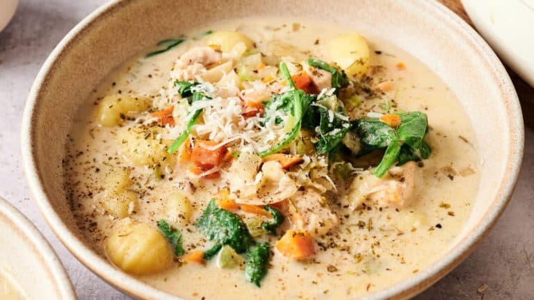 A bowl of creamy soup with gnocchi, spinach, carrots, chicken, and grated cheese, topped with herbs and black pepper.