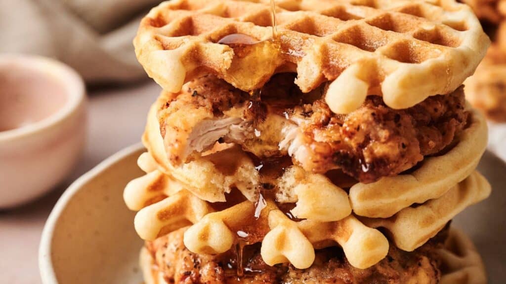 Stack of fried chicken and waffles with syrup being poured on top, served on a plate.