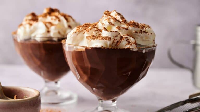 Two glass dessert cups filled with chocolate mousse, topped with whipped cream and chocolate shavings, on a light-colored surface.