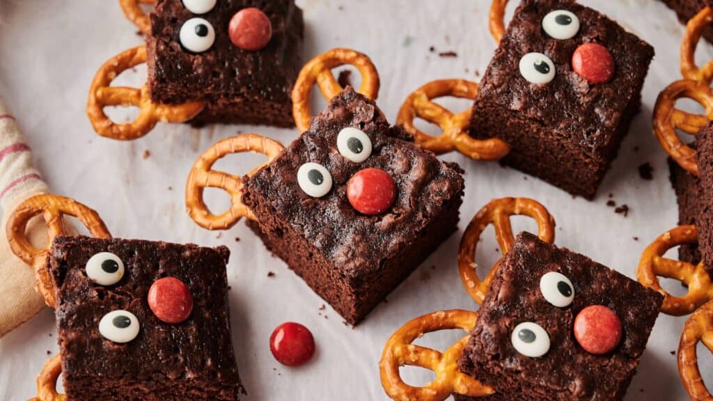 Brownie squares decorated with candy eyes, red candy noses, and pretzel antlers to resemble reindeer, arranged on a white surface.