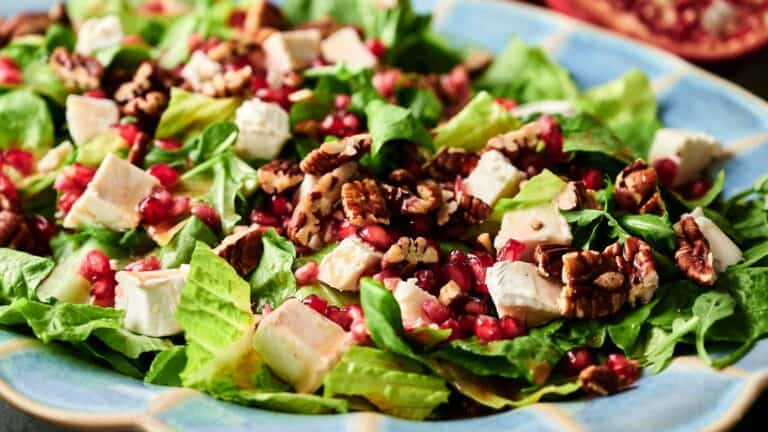 A salad with romaine lettuce, cubes of white cheese, pomegranate seeds, and chopped pecans on a blue plate.