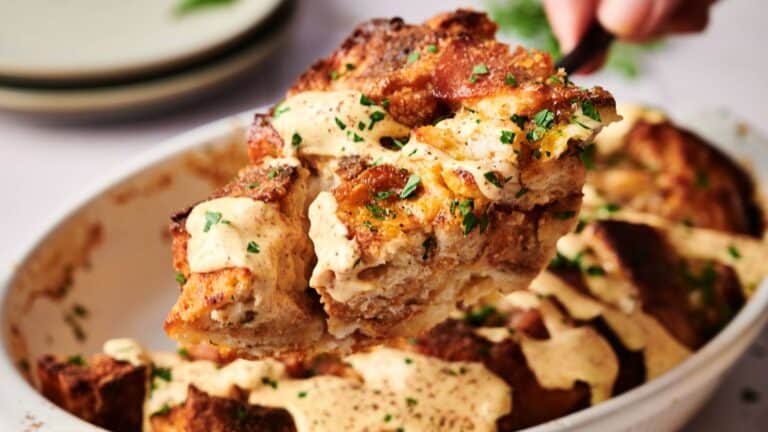 A close-up of a serving of bread pudding topped with creamy sauce and chopped herbs being lifted from a baking dish.