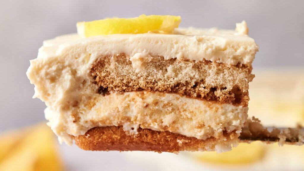 A close-up of a fork holding a slice of layered tiramisu dessert with creamy filling, biscuit layers, and a small lemon slice on top.