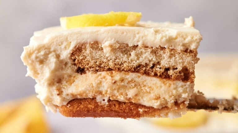 A close-up of a fork holding a slice of layered tiramisu dessert with creamy filling, biscuit layers, and a small lemon slice on top.