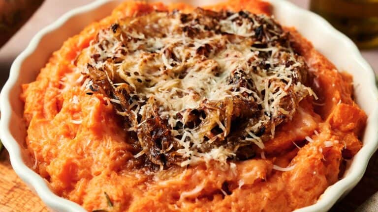 A white dish contains mashed sweet potatoes topped with caramelized onions and grated cheese.