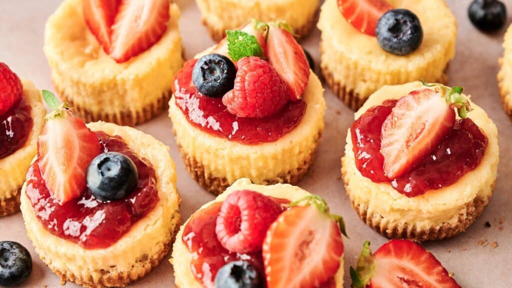 Mini cheesecakes topped with strawberry sauce, fresh strawberries, blueberries, and raspberries are arranged closely together on a light surface.