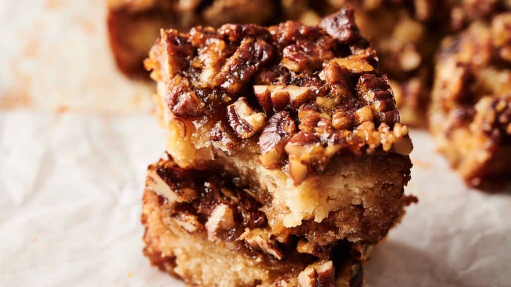 Two pecan bars stacked on top of each other, showing a nutty topping and a dense, golden-brown base on parchment paper.