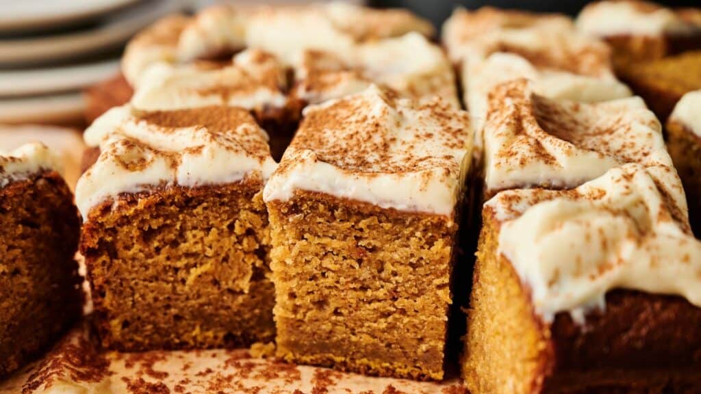 Close-up of sliced pumpkin bars topped with cream cheese frosting and a sprinkle of cinnamon, arranged on a parchment-lined surface.