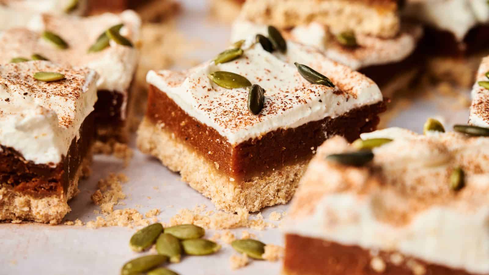Close-up of dessert bars with a crumbly base, pumpkin filling, whipped topping, dusted with cinnamon, and garnished with pumpkin seeds.