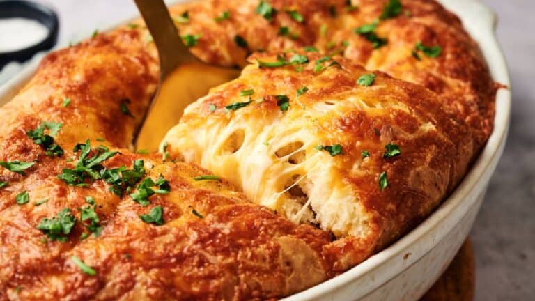 A close-up of a baked cheesy bread casserole, with a spoon lifting out a piece, topped with melted cheese and chopped parsley.