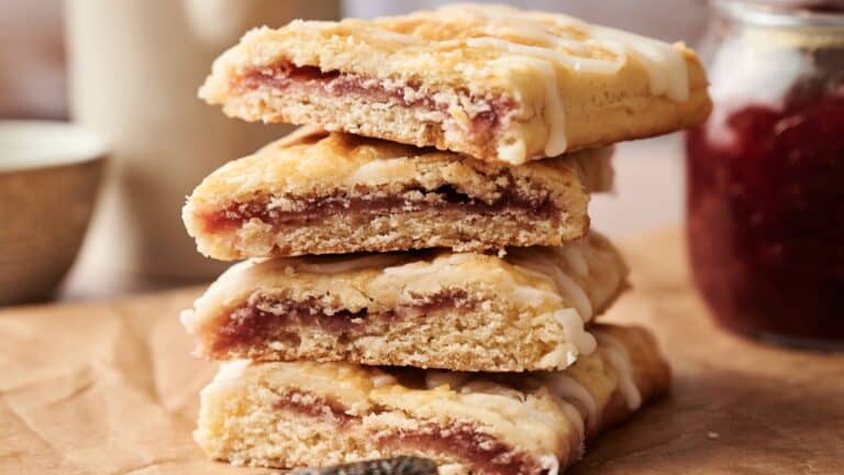 A stack of three rectangular pastries filled with red jam and drizzled with white icing, displayed on brown parchment paper.