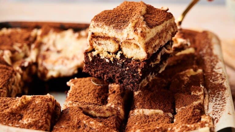 A close-up of a tray of tiramisu brownies, with one square being lifted, showing layers of chocolate brownie and cream, topped with cocoa powder.