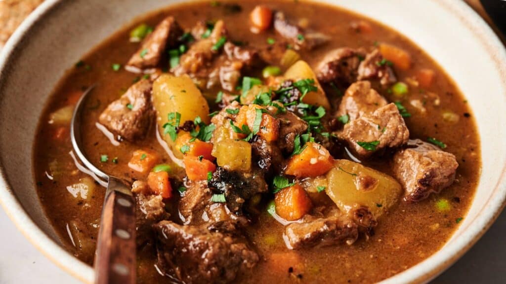 A bowl of beef stew with chunks of beef, potatoes, carrots, and peas in a brown broth, garnished with chopped herbs.
