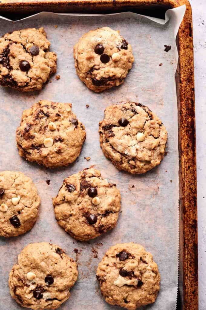 Seven Marry Me Cookies with visible white and dark chocolate chips are arranged on a parchment-lined, browned baking sheet.
