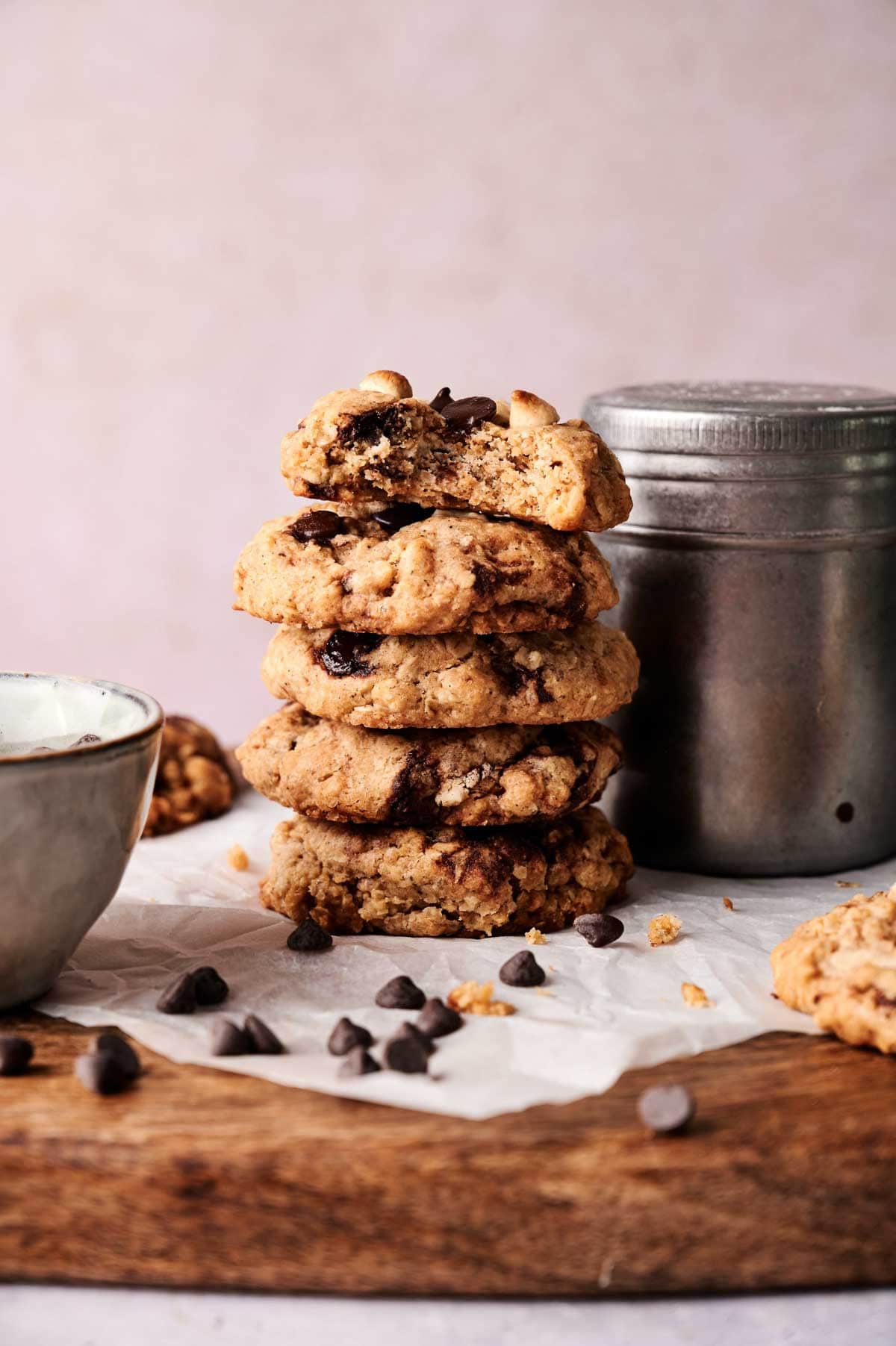 A stack of four Marry Me Cookies sits on parchment paper, with chocolate chips scattered around and a metal container and bowl nearby on a wooden surface.
