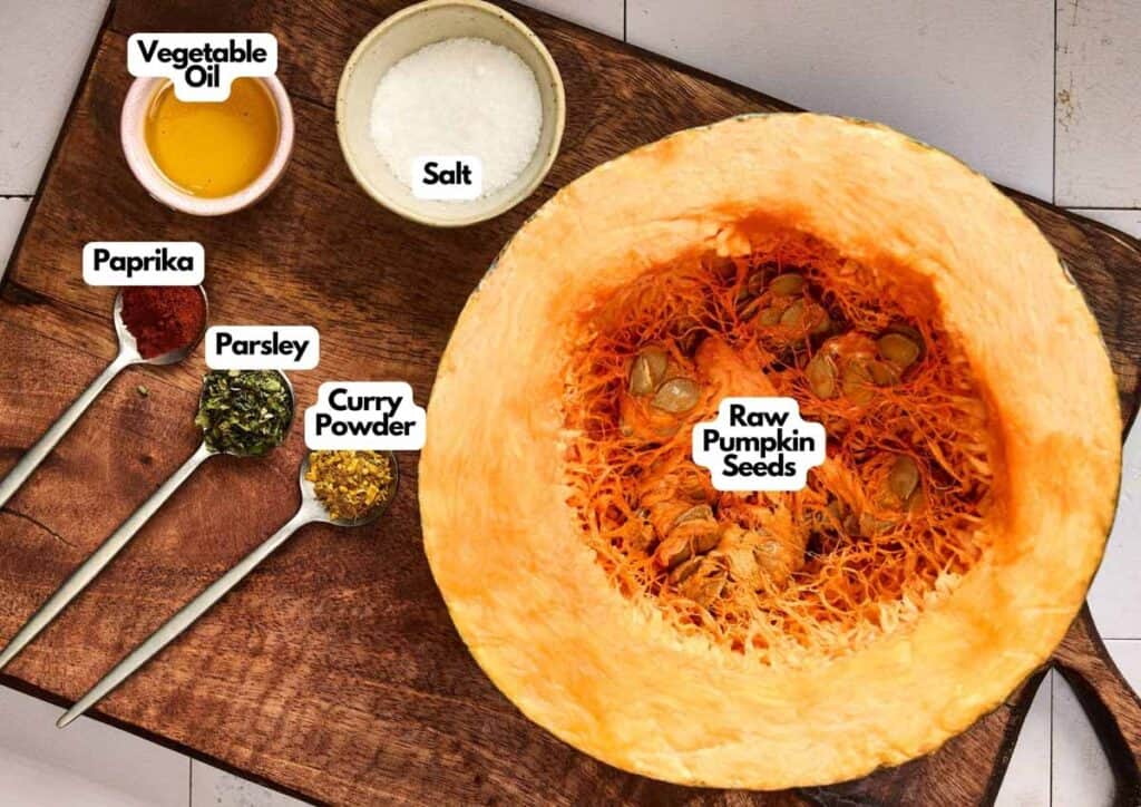A halved pumpkin with raw seeds, perfect for making Roasted Pumpkin Seeds, sits on a cutting board surrounded by labeled bowls of salt, vegetable oil, paprika, parsley, and curry powder.