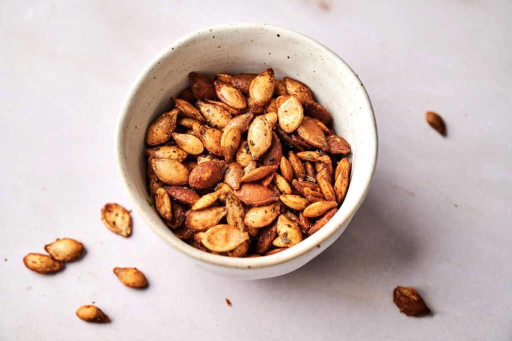 A white bowl filled with Roasted Pumpkin Seeds, perfectly seasoned, sits on a light surface with a few seeds scattered around it.