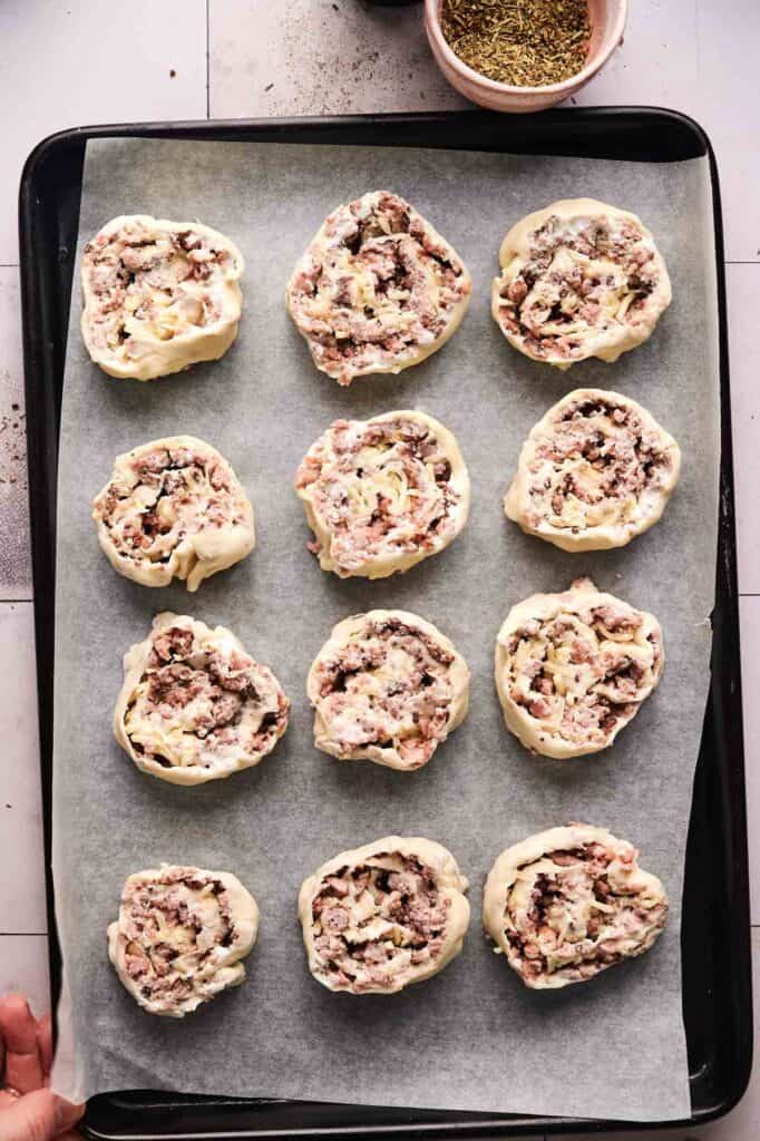 A baking tray lined with parchment paper holds twelve unbaked Sausage Pinwheels, each pastry roll filled with a savory minced meat and onion mixture.