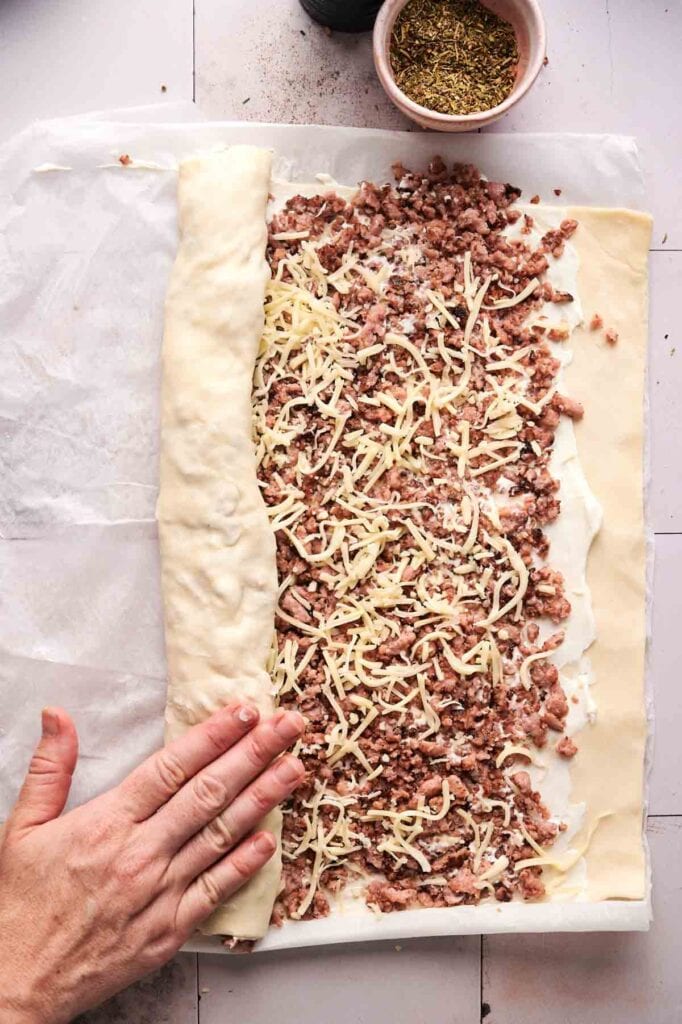 A hand rolls up dough filled with ground meat and shredded cheese on parchment paper, preparing delicious Sausage Pinwheels, with a bowl of dried herbs nearby.