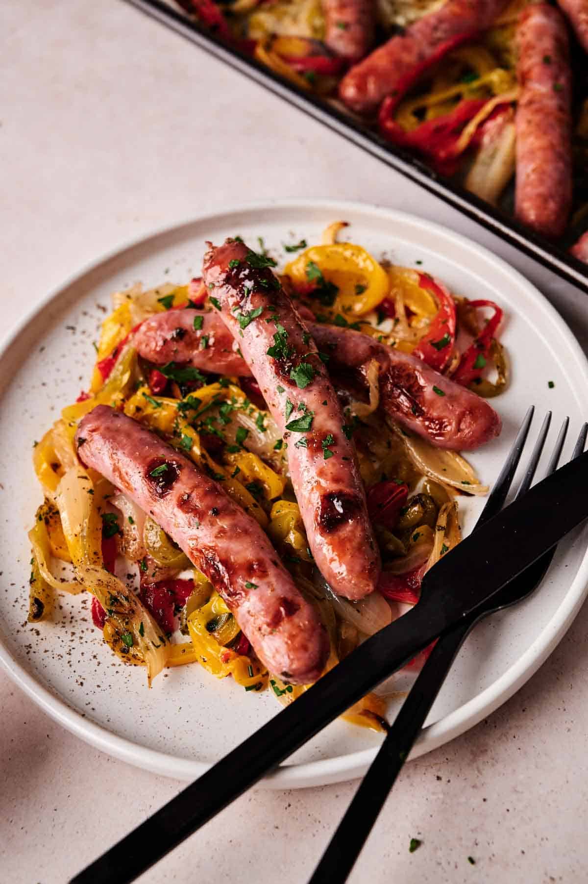 Three grilled sausages served over saut&eacute;ed bell peppers and onions, garnished with chopped herbs&mdash;this Sheet Pan Sausage and Peppers dish is plated on white, alongside a black fork and knife.