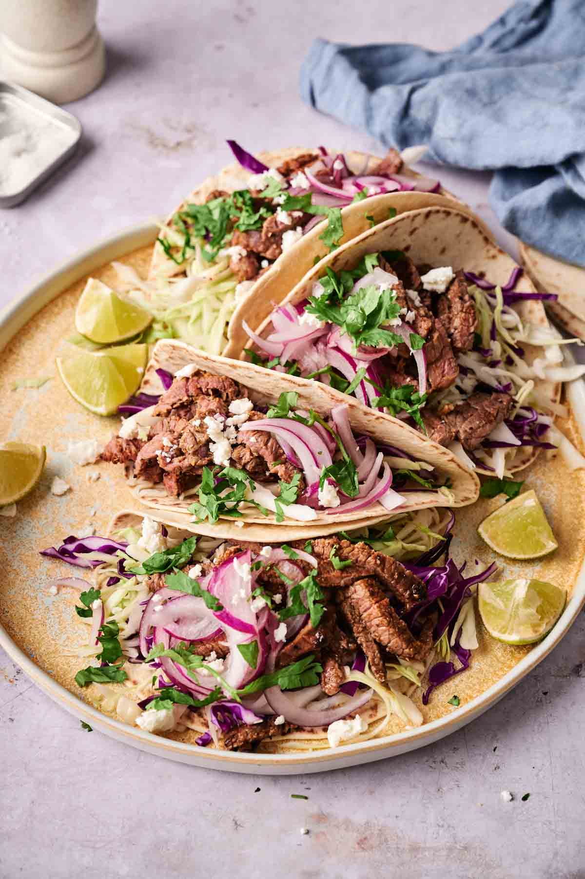 Three Street Steak Tacos on a plate, topped with shredded cabbage, red onion, cilantro, and crumbled cheese, served with lime wedges on the side.