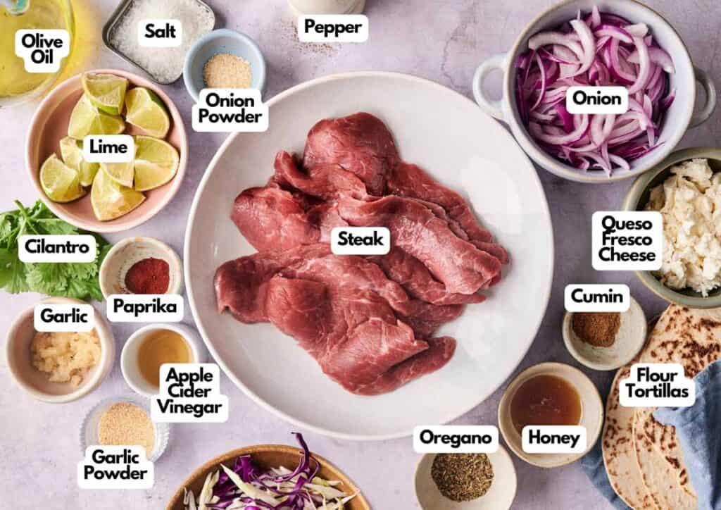 A bowl of raw steak for Street Steak Tacos is surrounded by labeled ingredients like onion, lime, spices, cheese, garlic, olive oil, tortillas, and seasonings on a white surface.