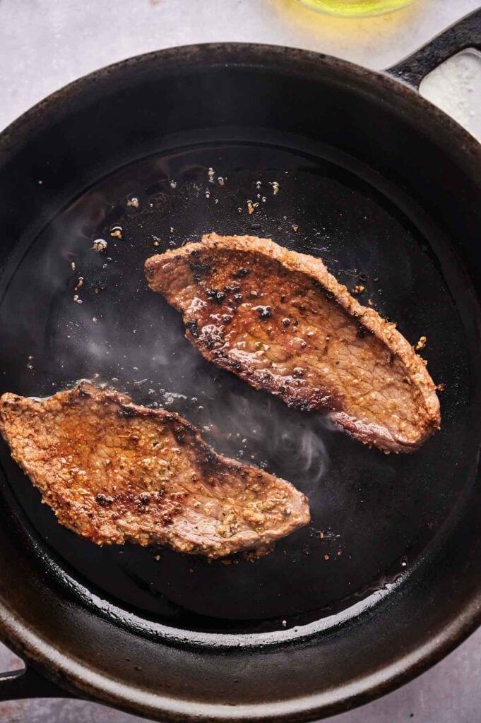 Two pieces of seasoned steak sizzle in a cast iron skillet, with steam rising from the meat&mdash;perfect for making delicious Street Steak Tacos.
