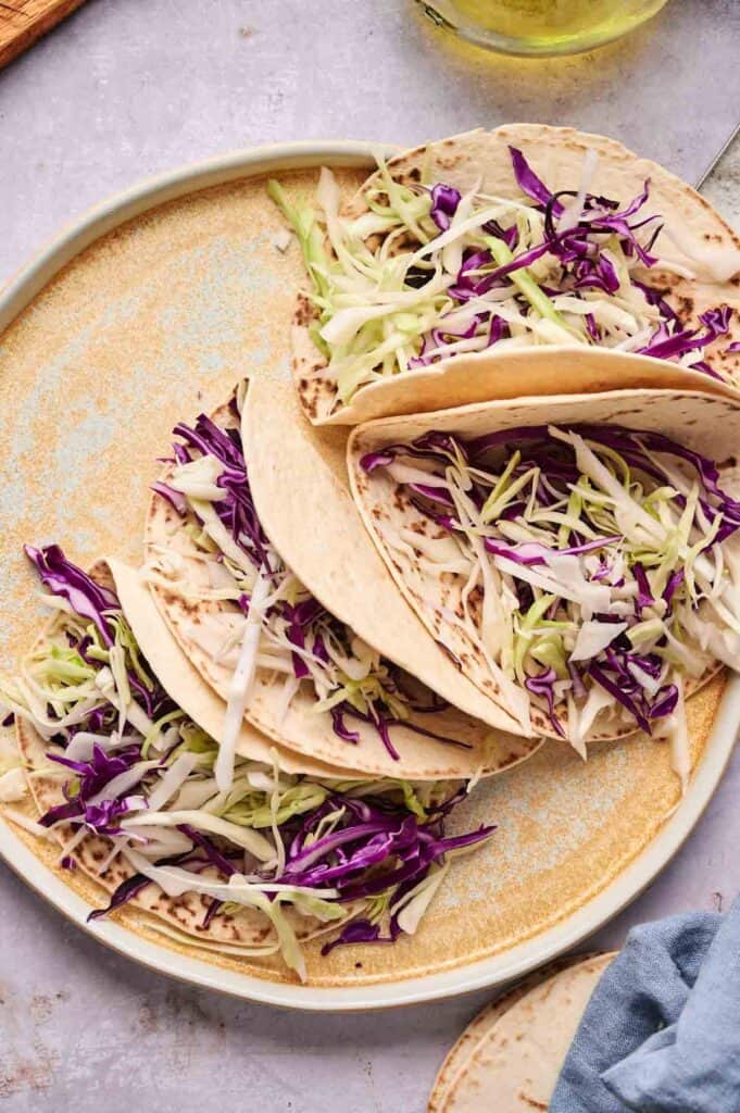 Three soft Street Steak Tacos on a plate, filled with shredded green and purple cabbage, with no other visible toppings.