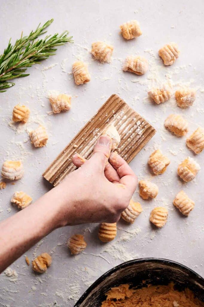A hand shapes sweet potato gnocchi on a wooden board, with pieces of uncooked gnocchi and a sprig of rosemary scattered on the floured surface.