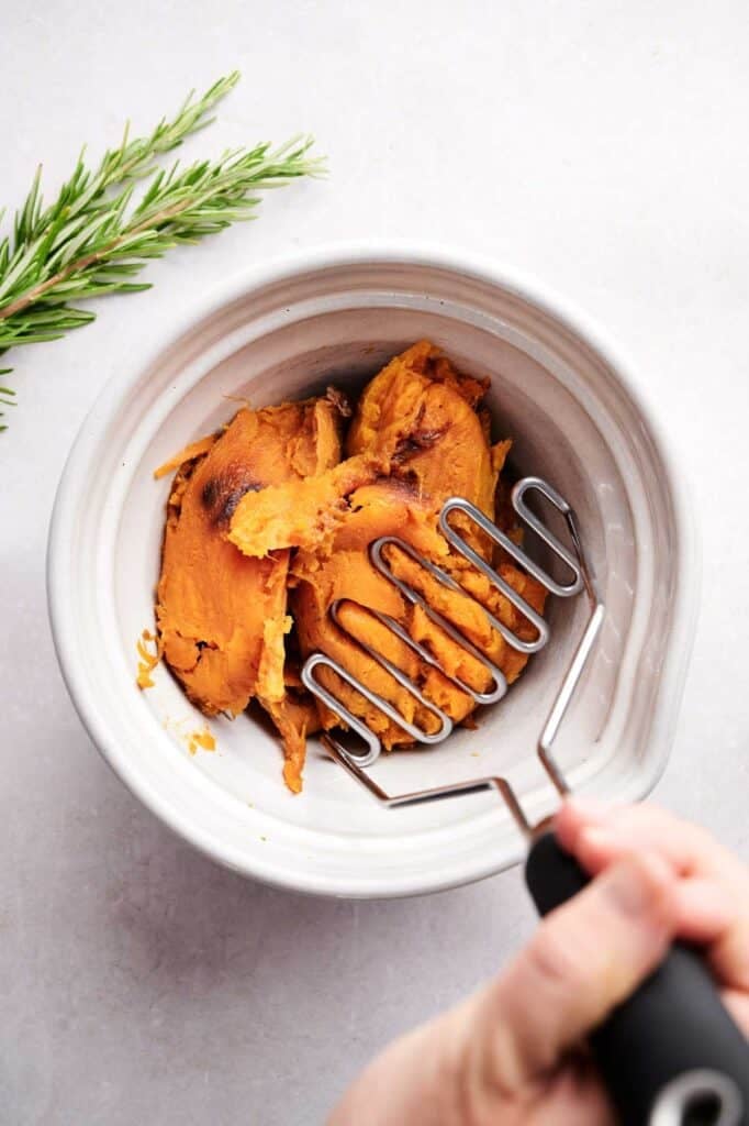 A hand holds a potato masher over cooked sweet potatoes in a white bowl, ready to start making sweet potato gnocchi, with a sprig of rosemary nearby on a light surface.