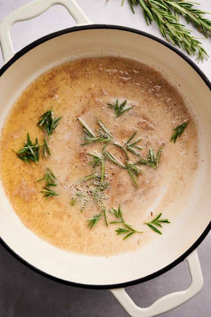 A white pot with browned butter and rosemary sprigs on a light surface, perfect for tossing Sweet Potato Gnocchi, with extra rosemary in the background.