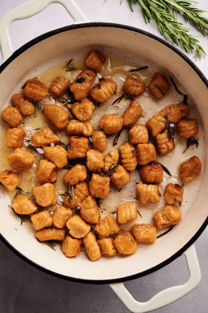 Pan-fried Sweet Potato Gnocchi with fresh rosemary in a white cast iron skillet, surrounded by a few sprigs of rosemary on a light surface.