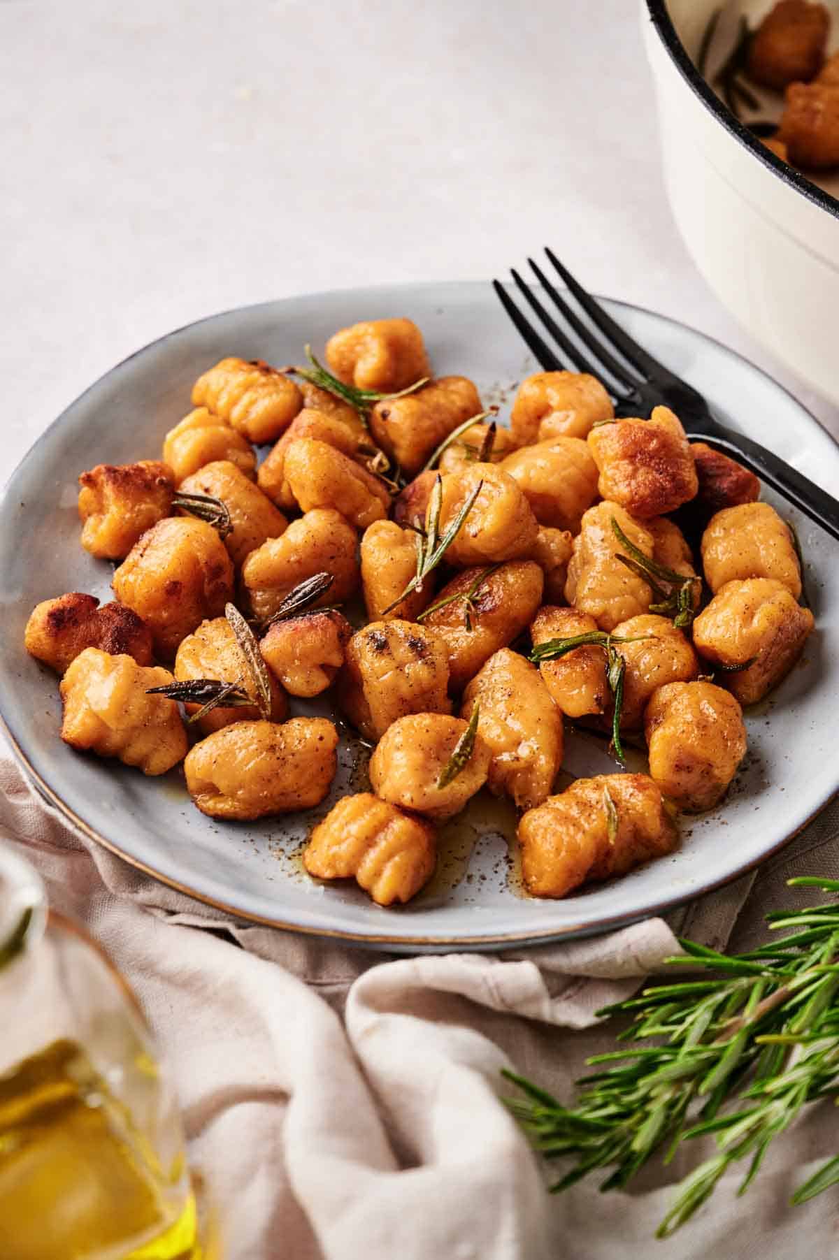 A plate of browned sweet potato gnocchi garnished with rosemary, with two black forks on the side. A sprig of fresh rosemary and a glass bottle of olive oil are nearby.