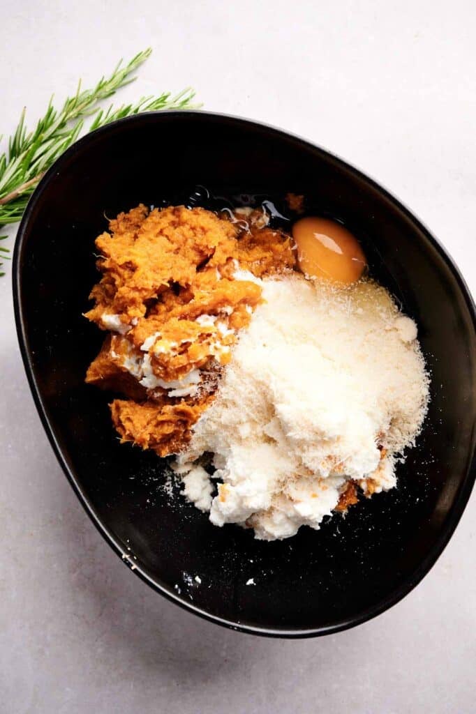 A black bowl containing sweet potato puree, ricotta cheese, an egg, grated cheese, and a sprig of rosemary on the side—perfect ingredients for homemade Sweet Potato Gnocchi.