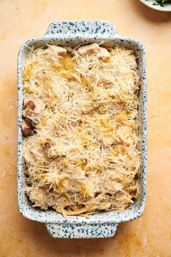 A rectangular baking dish filled with uncooked Turkey Tetrazzini casserole, topped with shredded cheese and breadcrumbs, rests on a yellow surface.