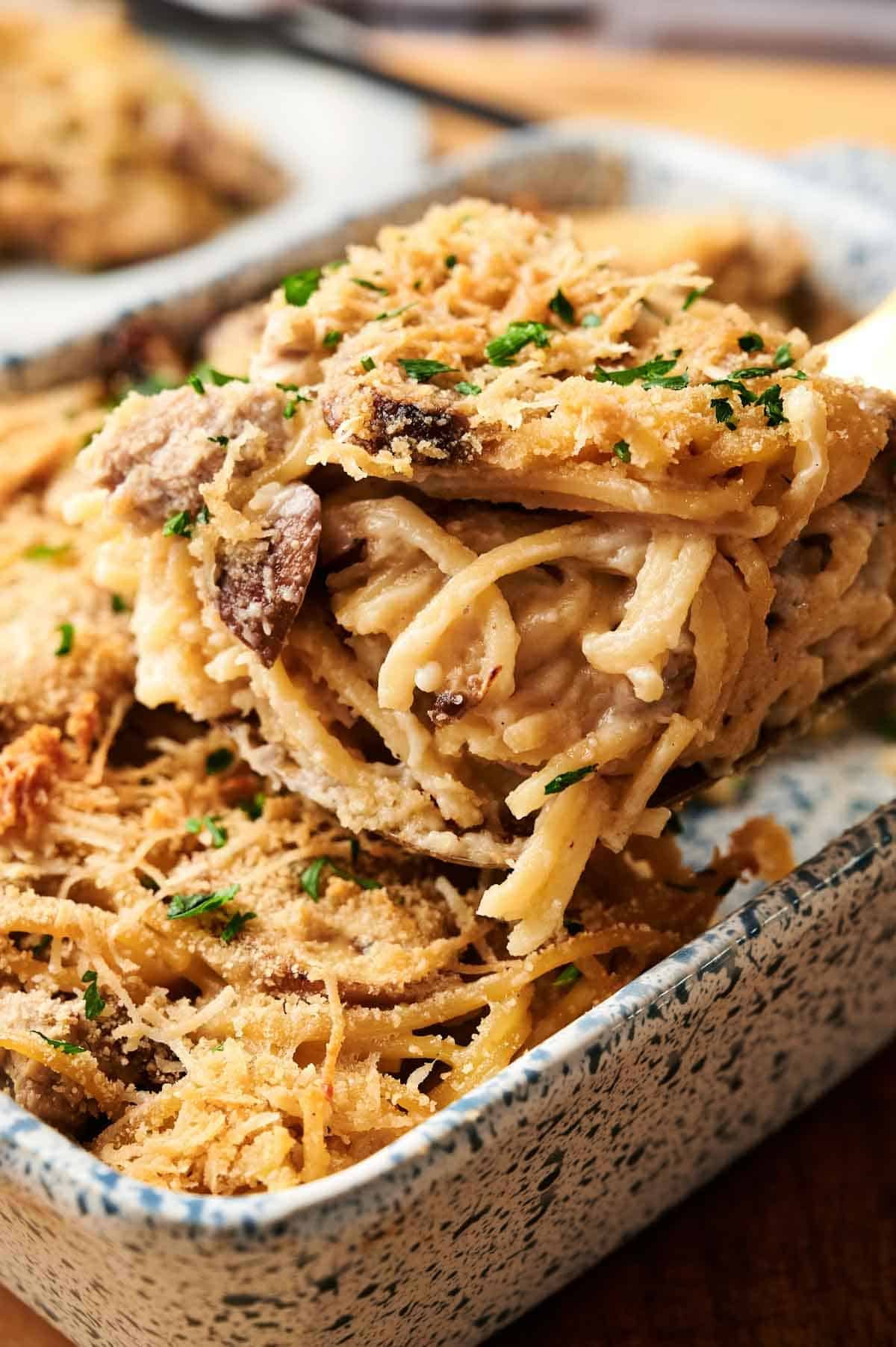 Close-up of a serving of Turkey Tetrazzini baked with mushrooms and a golden, crispy cheese topping, garnished with chopped parsley in a speckled baking dish.