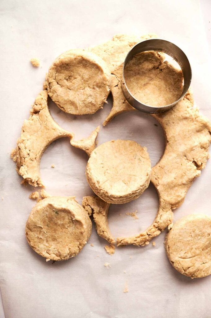 A round metal biscuit cutter cutting circles from rolled-out Cheddar Biscuits dough on parchment paper, with several biscuit shapes and dough scraps visible.