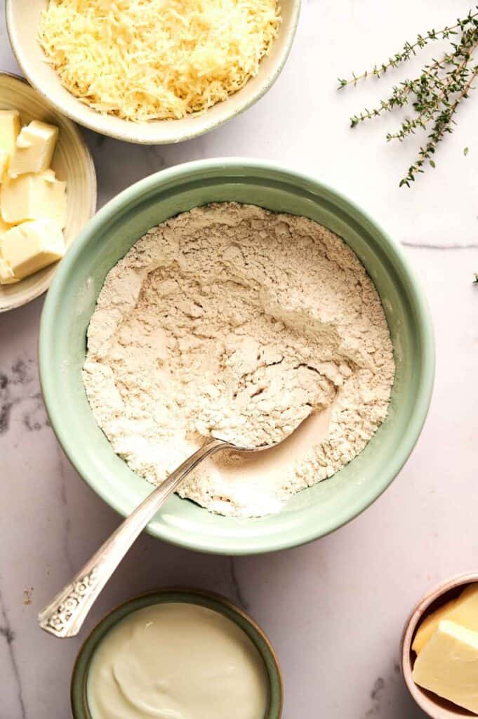 A bowl of flour with a spoon, surrounded by bowls of grated cheese, butter, and a small bowl of cream on a marble surface with a sprig of thyme—perfect ingredients for making Cheddar Biscuits.