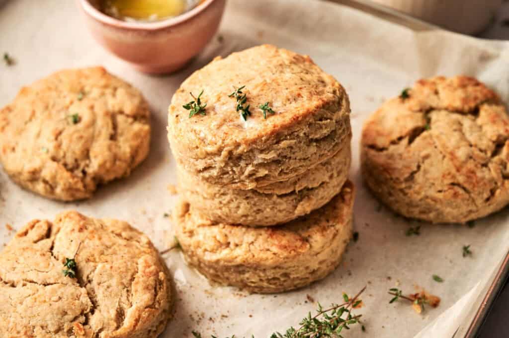 Four golden brown Cheddar Biscuits with a flaky texture are stacked on parchment paper, garnished with small sprigs of thyme; a bowl of melted butter is in the background.