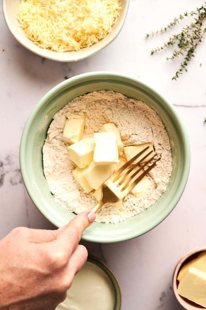 A hand holds a fork, mixing cubes of butter into flour for Cheddar Biscuits. Grated cheese, fresh herbs, and a bowl of milk are nearby on a white surface.