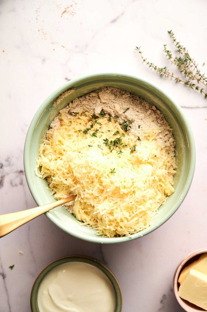 A green bowl filled with grated cheese, flour, and fresh thyme—perfect for making Cheddar Biscuits—sits on a marble surface with a gold spoon inside.