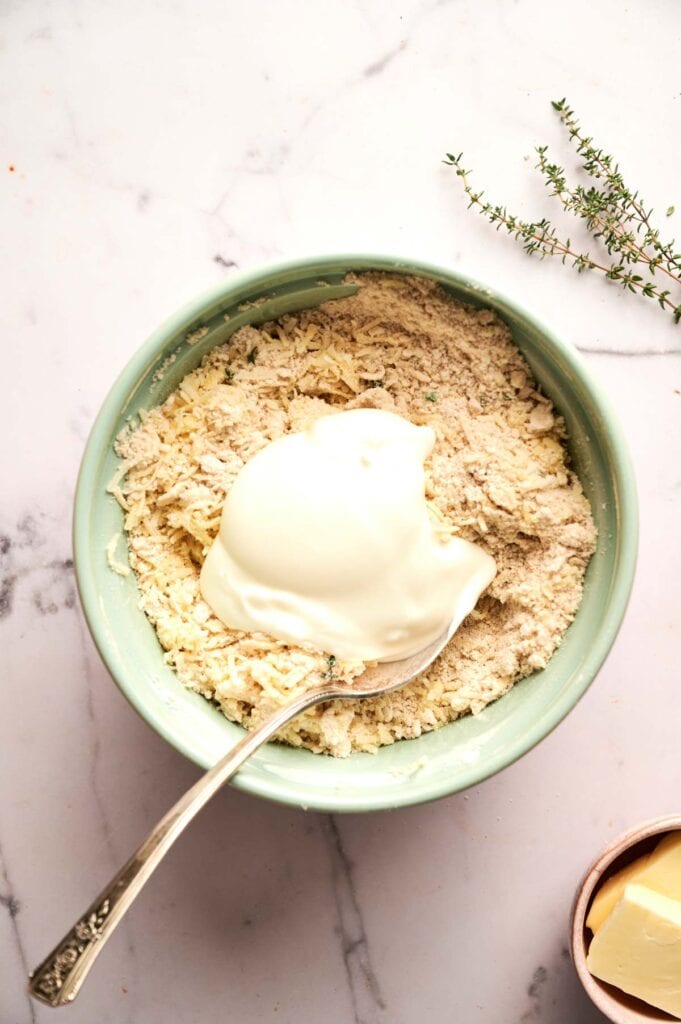 A green bowl holds flour mixture for Cheddar Biscuits, topped with a large spoonful of sour cream; a sprig of thyme and a butter pat rest nearby on the marble surface.
