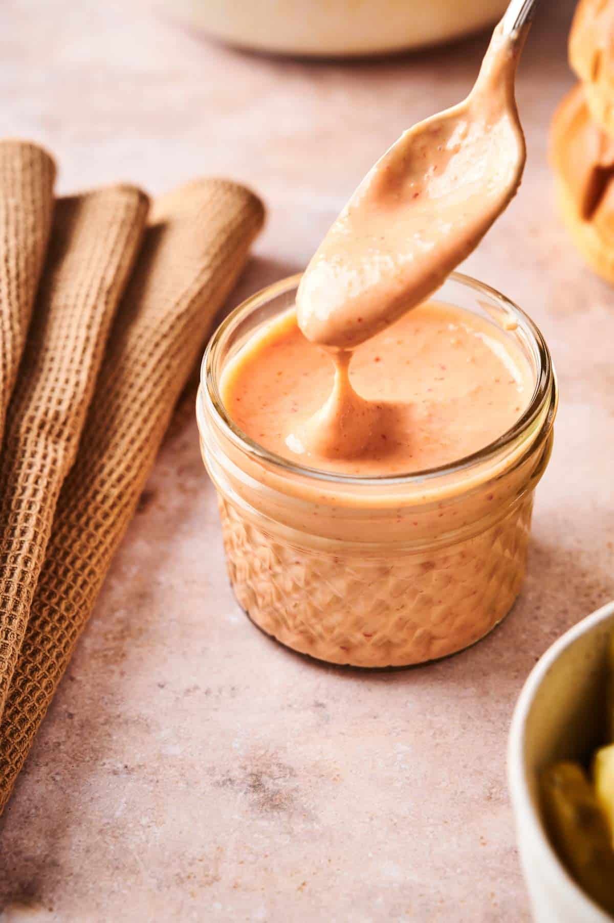 A spoon is held over a small glass jar filled with creamy, orange-colored Boom Boom Sauce on a light countertop, next to a folded brown cloth.